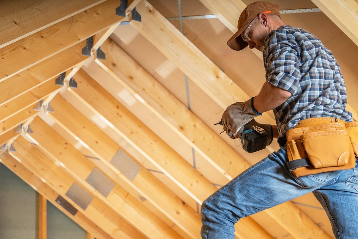 A construction worker is focused on installing wooden beams in an attic space during a home renovation. Visible are the wooden structures overhead, showcasing the progress of the project in bright, natural light.