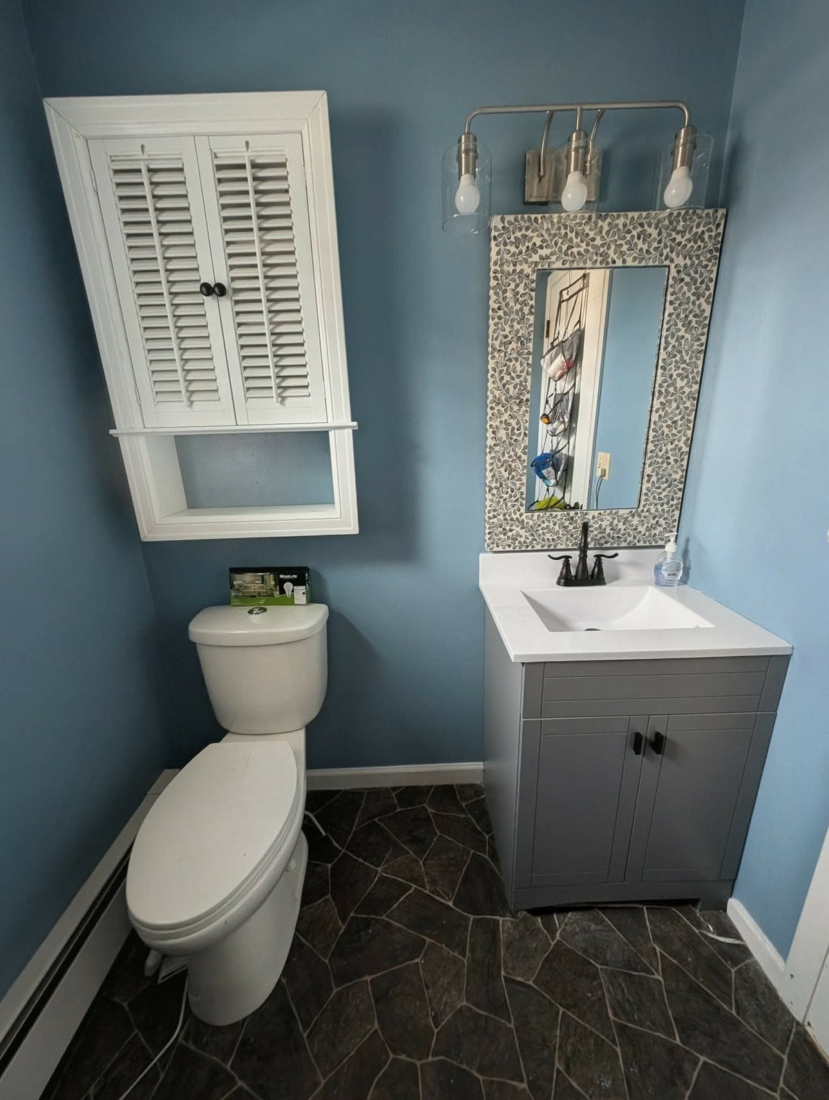 Modern blue bathroom with white toilet, gray vanity sink, decorative mirror, and white shutter cabinet above