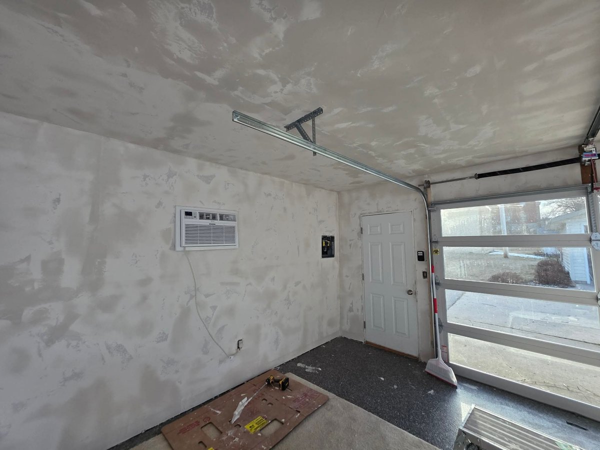 Unfinished garage interior with drywall, white door, open garage door, window, and floor materials