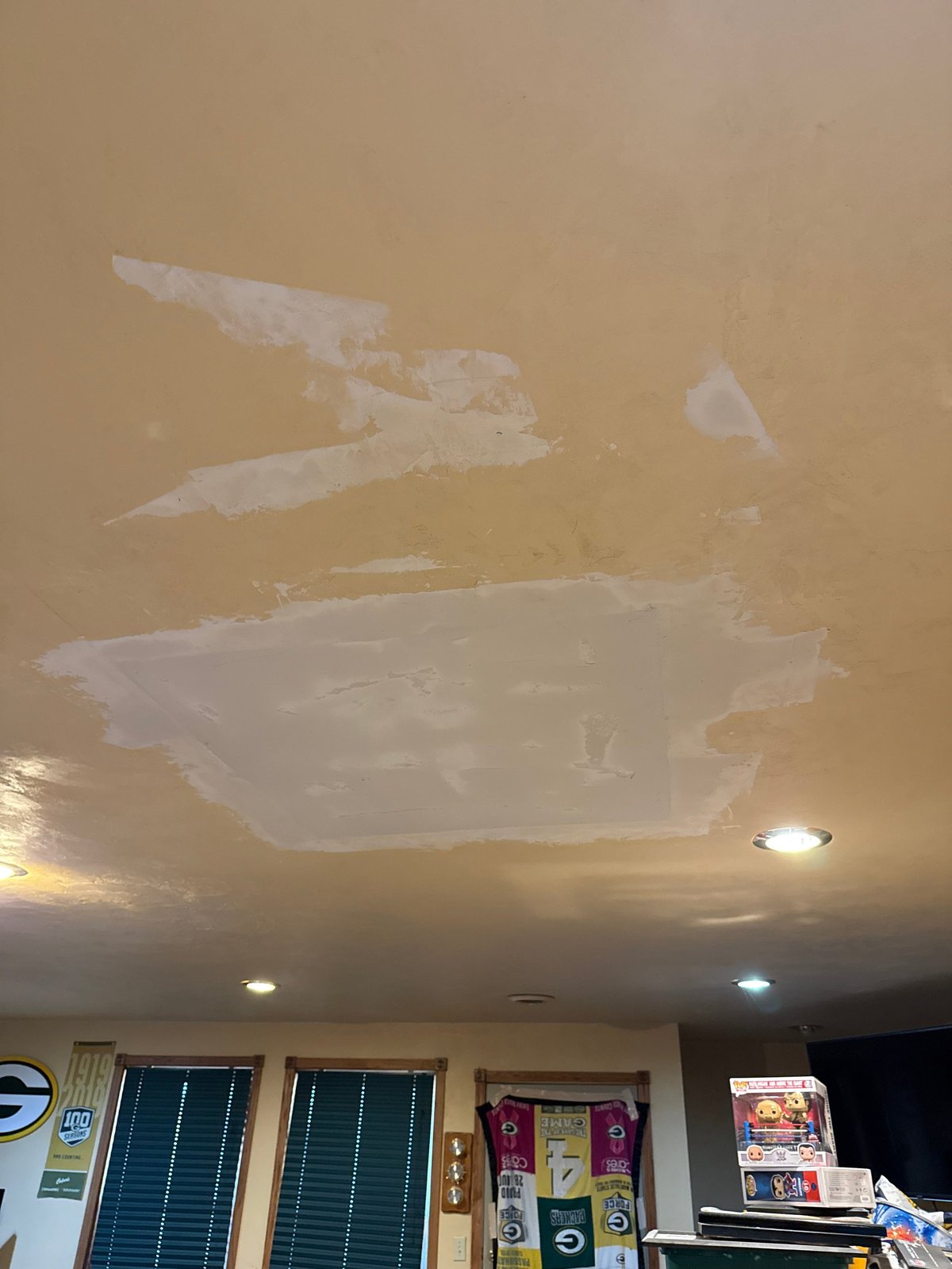 Ceiling with multiple patched drywall repairs and recessed lighting in a residential room with windows and arcade game visible