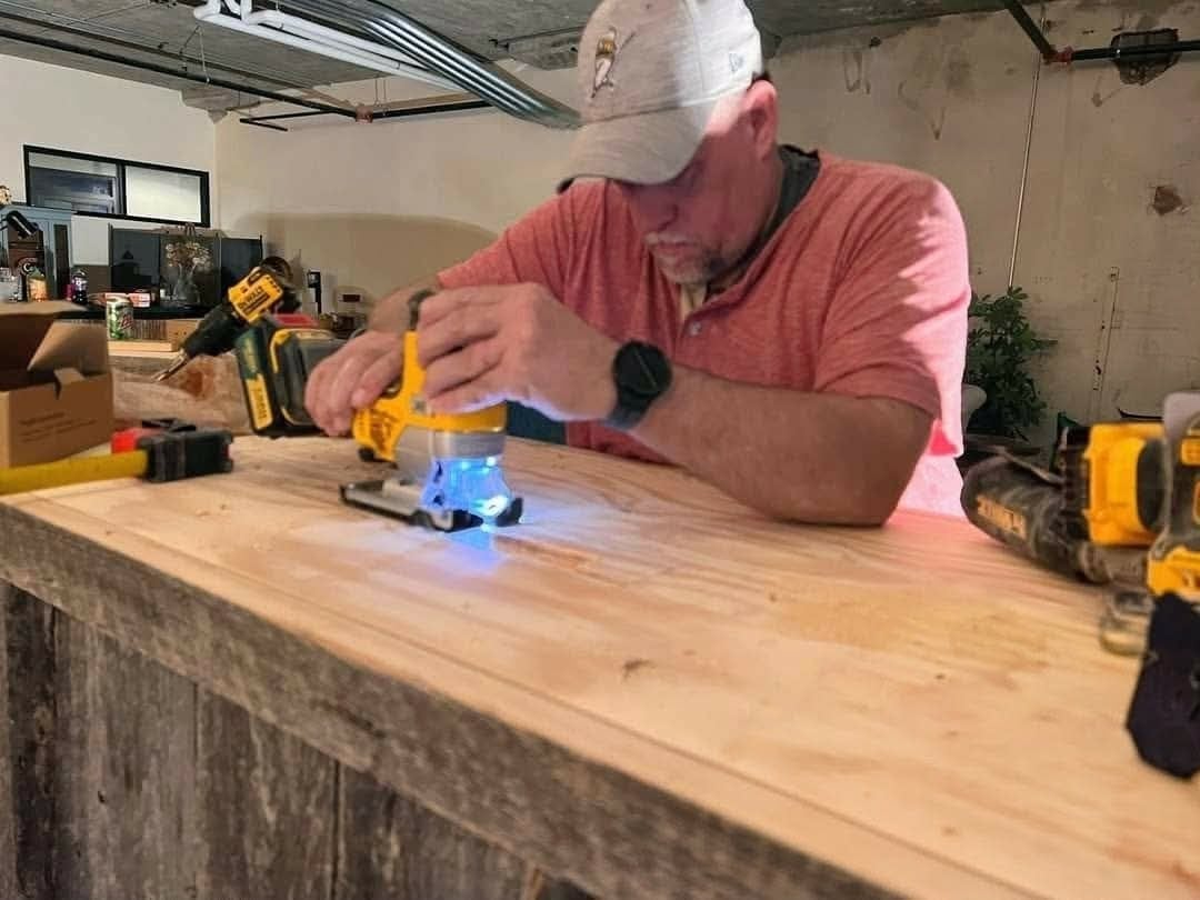 Man in red shirt and white cap demonstrates a yellow power tool on wooden workbench in workshop setting