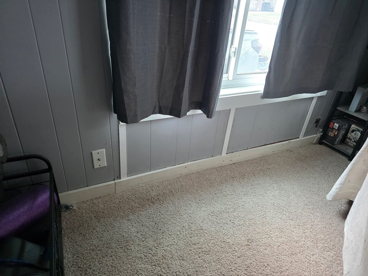 Small bedroom with beige carpet, gray walls, dark curtains over window, white wainscoting, and minimal furniture