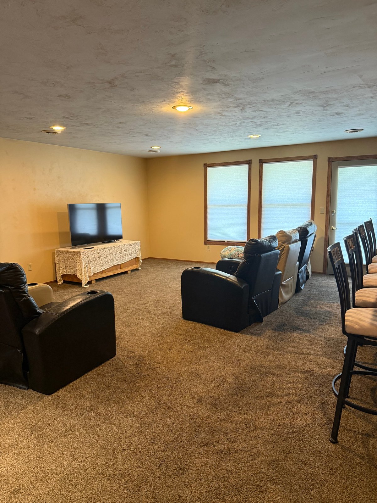 Spacious basement recreation room with yellow walls, large windows, black armchairs, wicker TV stand, and bar seating area
