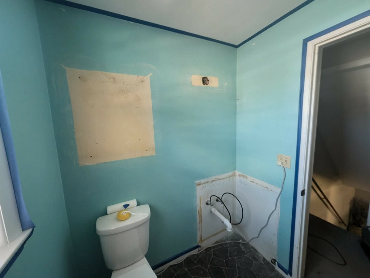 A bathroom under renovation with turquoise walls, white toilet, uncovered drywall patches, and exposed wiring