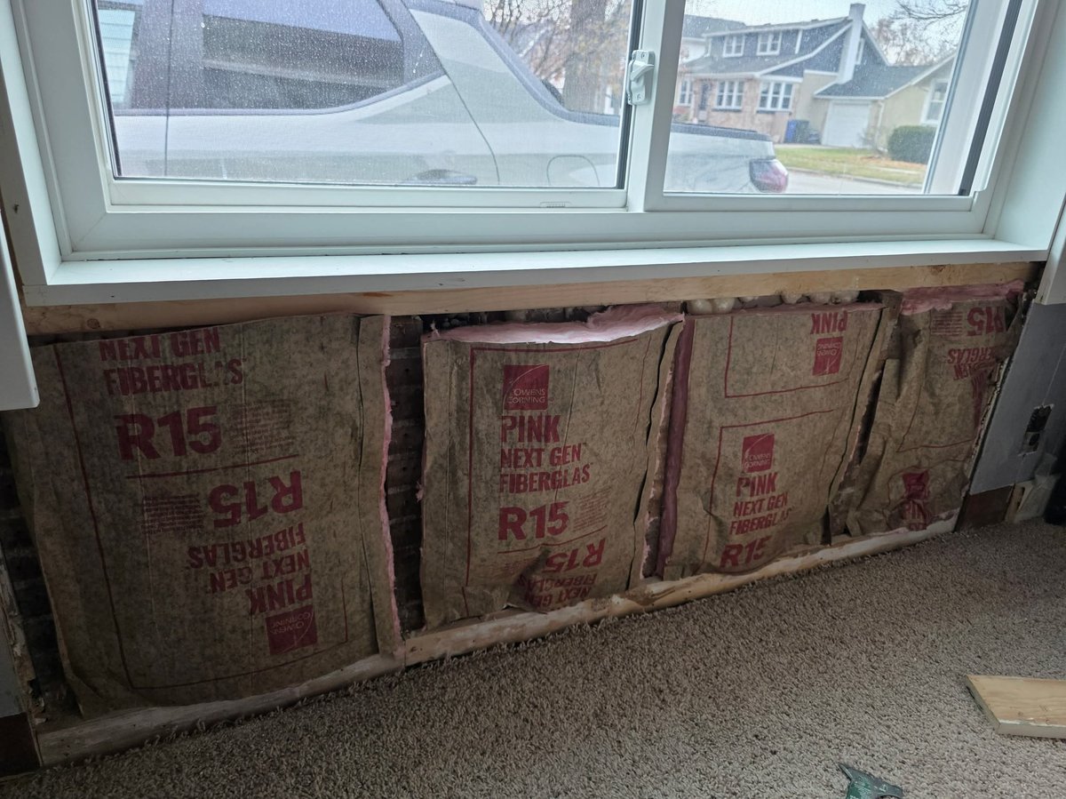 Three insulation batts wrapped in kraft paper labeled R15 stacked under a window in a room under construction